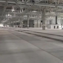 Wnętrze hali produkcyjnej w trakcie budowy, widoczne szyny w posadzce, stalowa konstrukcja dachu i słupy nośne, pomarańczowy podnośnik koszowy w tle.