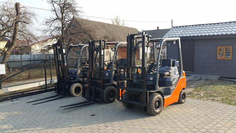 Trzy wózki widłowe marki Toyota, jeden pomarańczowy, dwa szare, zaparkowane na brukowanym placu przed szarym budynkiem z drewnianą elewacją.