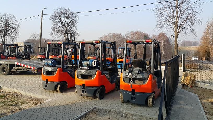 Rząd nowych wózków widłowych marki Toyota w kolorze pomarańczowo-szarym, zaparkowanych na placu z kostki brukowej, z przyczepą transportową i budynkami w tle.