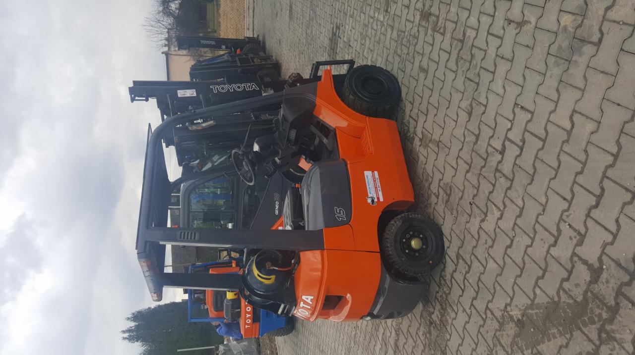 Pomarańczowy wózek widłowy Toyota GENEO 20 z numerem 15 na boku, stojący na brukowanym placu, z innymi wózkami widłowymi w tle.