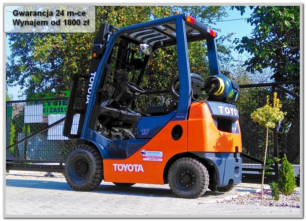 Nowy wózek widłowy Toyota, pomarańczowo-niebieski, z naklejką informującą o gwarancji i możliwości wynajmu, zaparkowany na zewnątrz z widocznym ogrodzeniem i zielenią w tle.