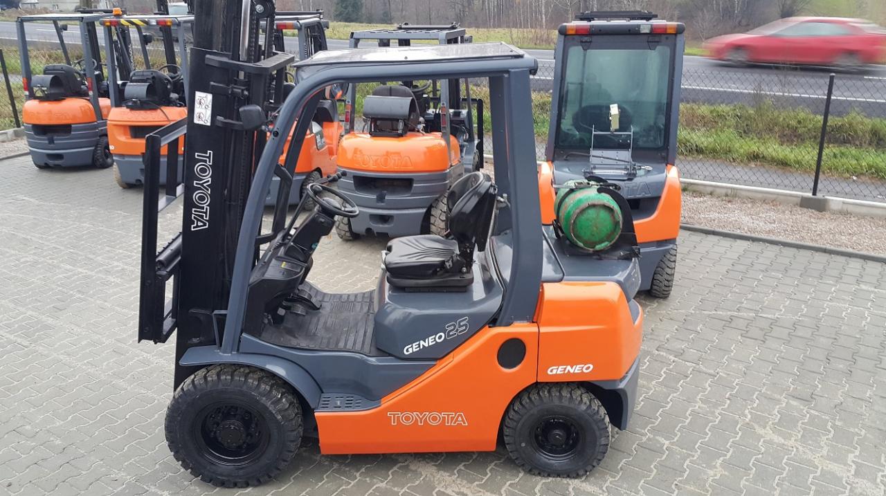 Pomarańczowo-szary wózek widłowy Toyota Geneo25 na kostce brukowej, z butlą gazową z tyłu. W tle inne wózki i przejeżdżający samochód, co dodaje dynamiki.