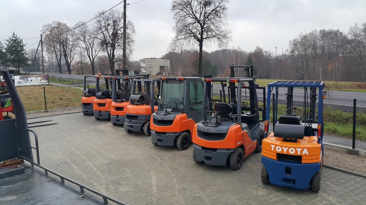 Rząd nowych wózków widłowych Toyota w kolorach pomarańczowym i szarym, stojących na placu w pochmurny dzień, z widoczną drogą w tle.