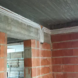 Surowe wnętrze w trakcie budowy z widocznymi przewodami elektrycznymi ułożonymi pod sufitem i wzdłuż ścian z czerwonej cegły.