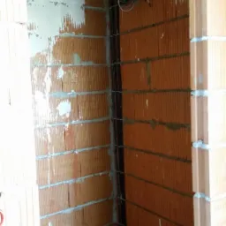 Wewnętrzny róg pomieszczenia w trakcie budowy z widocznymi przewodami elektrycznymi w bruzdach w ceglanej ścianie oraz zwisającym kablem.