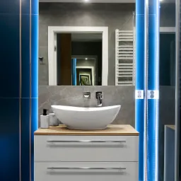 Nowoczesna łazienka z białą umywalką nablatową, szafką z dwiema szufladami na drewnianym blacie, szarymi płytkami na ścianie i podświetleniem LED wzdłuż krawędzi lustra i ściany.