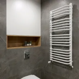 Nowoczesna łazienka z szarymi płytkami imitującymi marmur, podwieszana toaleta, biały grzejnik drabinkowy i szafka z białą zabudową oraz drewnianą wnęką z dekoracyjnymi świecami.