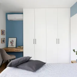 Nowoczesna sypialnia z białą zabudową szaf, biurkiem i niebieską ścianą, widok z perspektywy łóżka w paski.