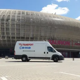 Biały bus firmy MT-Transport z numerem telefonu 691 242 691 zaparkowany na parkingu przed Tauron Areną w Krakowie, widoczny srebrny samochód osobowy.