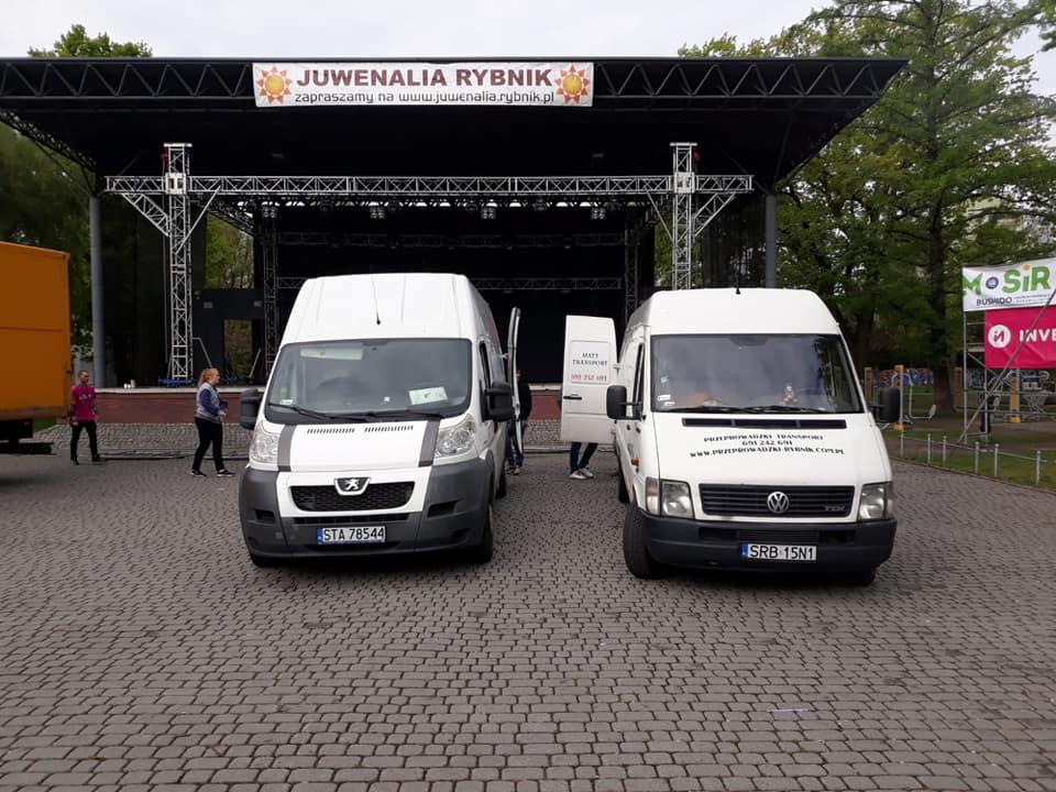 Dwa białe busy dostawcze na brukowanym placu przed sceną Juwenaliów Rybnik z otwartymi drzwiami, widoczny baner z informacjami kontaktowymi na jednym z busów.