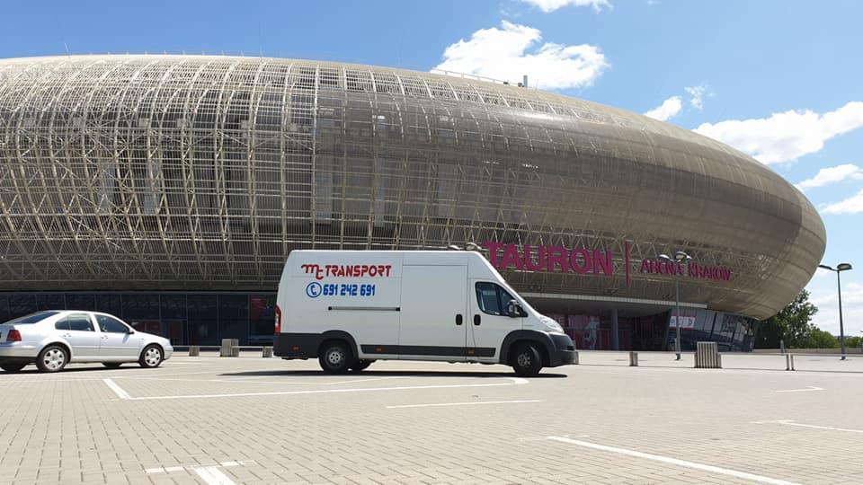 Biały bus firmy MT-Transport z numerem telefonu 691 242 691 zaparkowany na parkingu przed Tauron Areną w Krakowie, widoczny srebrny samochód osobowy.