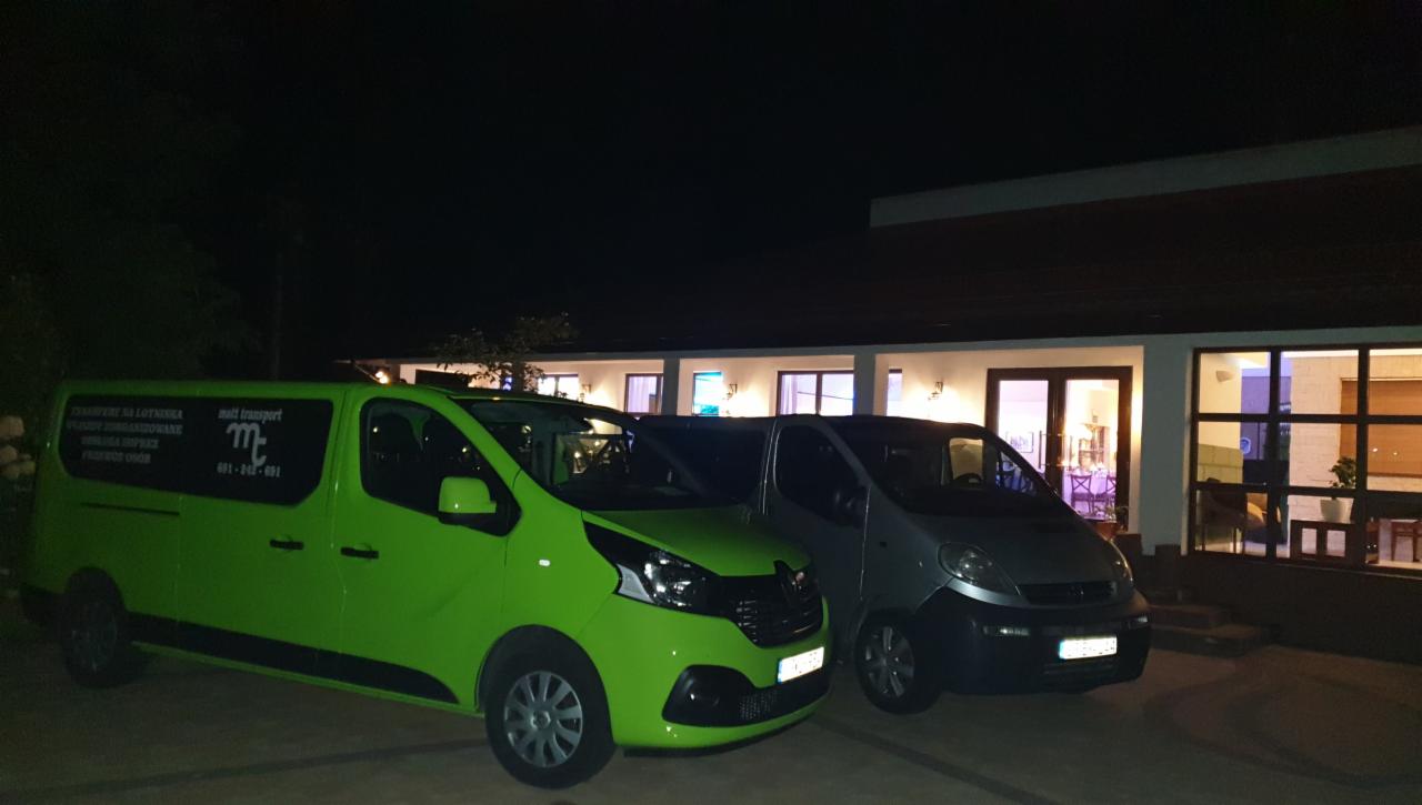 Dwa busy transportowe, jeden jaskrawo zielony z logo firmy i tekstem o transporcie na bocznej szybie, zaparkowane przed budynkiem z oświetlonymi oknami nocą.