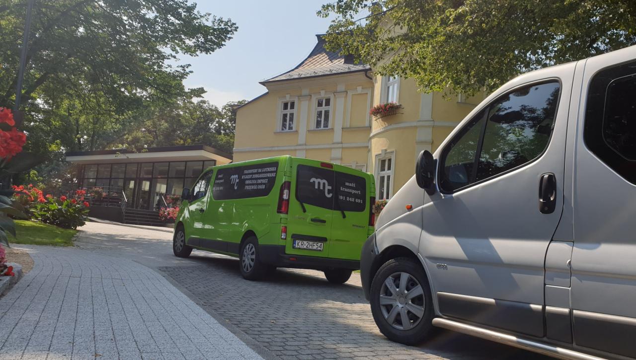 Dwa busy zaparkowane na brukowanej drodze przed budynkiem z czerwonymi kwiatami. Zielony bus z logo firmy transportowej i numerem telefonu na tylnej klapie.