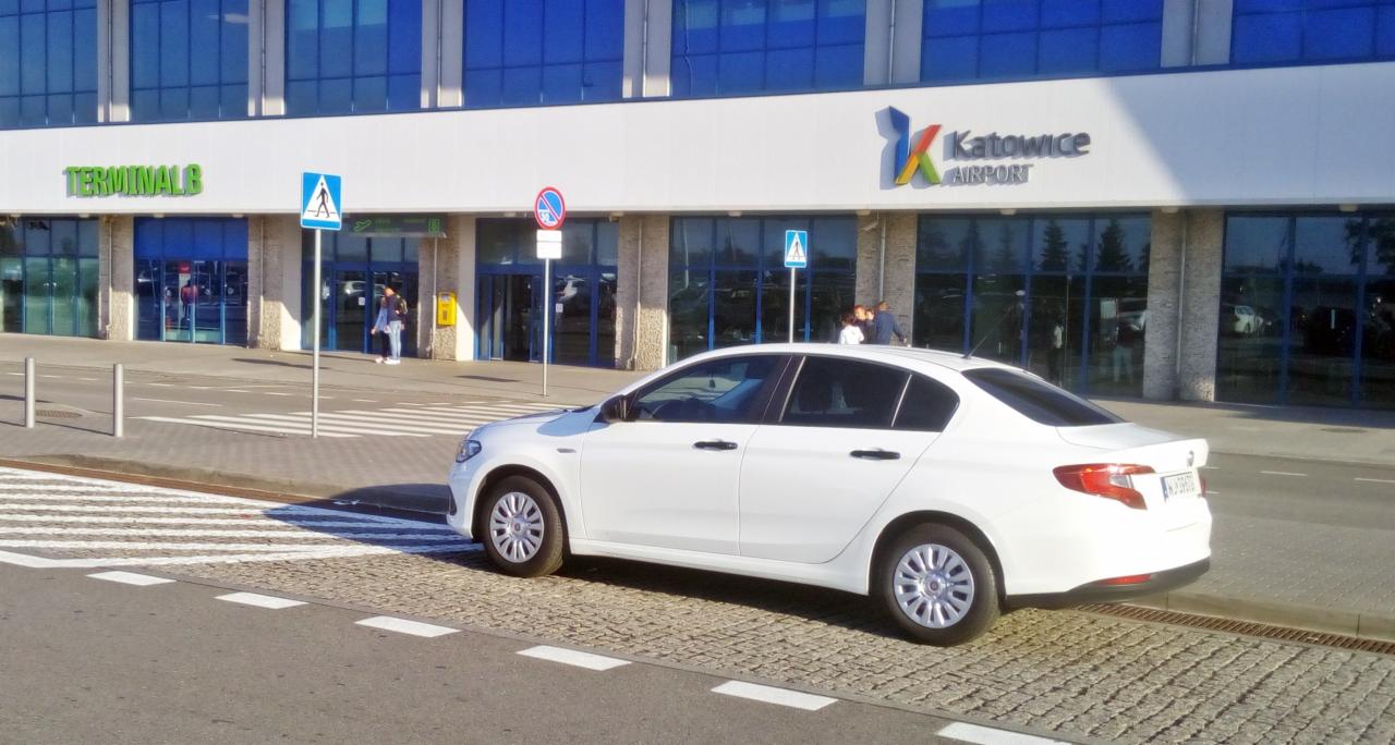 Biały samochód osobowy na przejściu dla pieszych przed budynkiem lotniska Katowice Airport w słoneczny dzień.