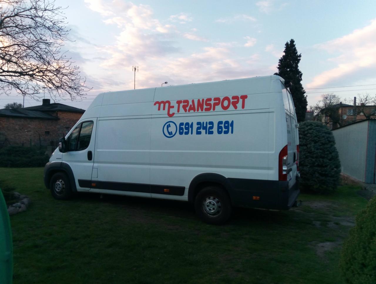Biały samochód dostawczy z czerwonym napisem 'm-TRANSPORT' i niebieskim numerem telefonu na tle zielonego trawnika i budynków.