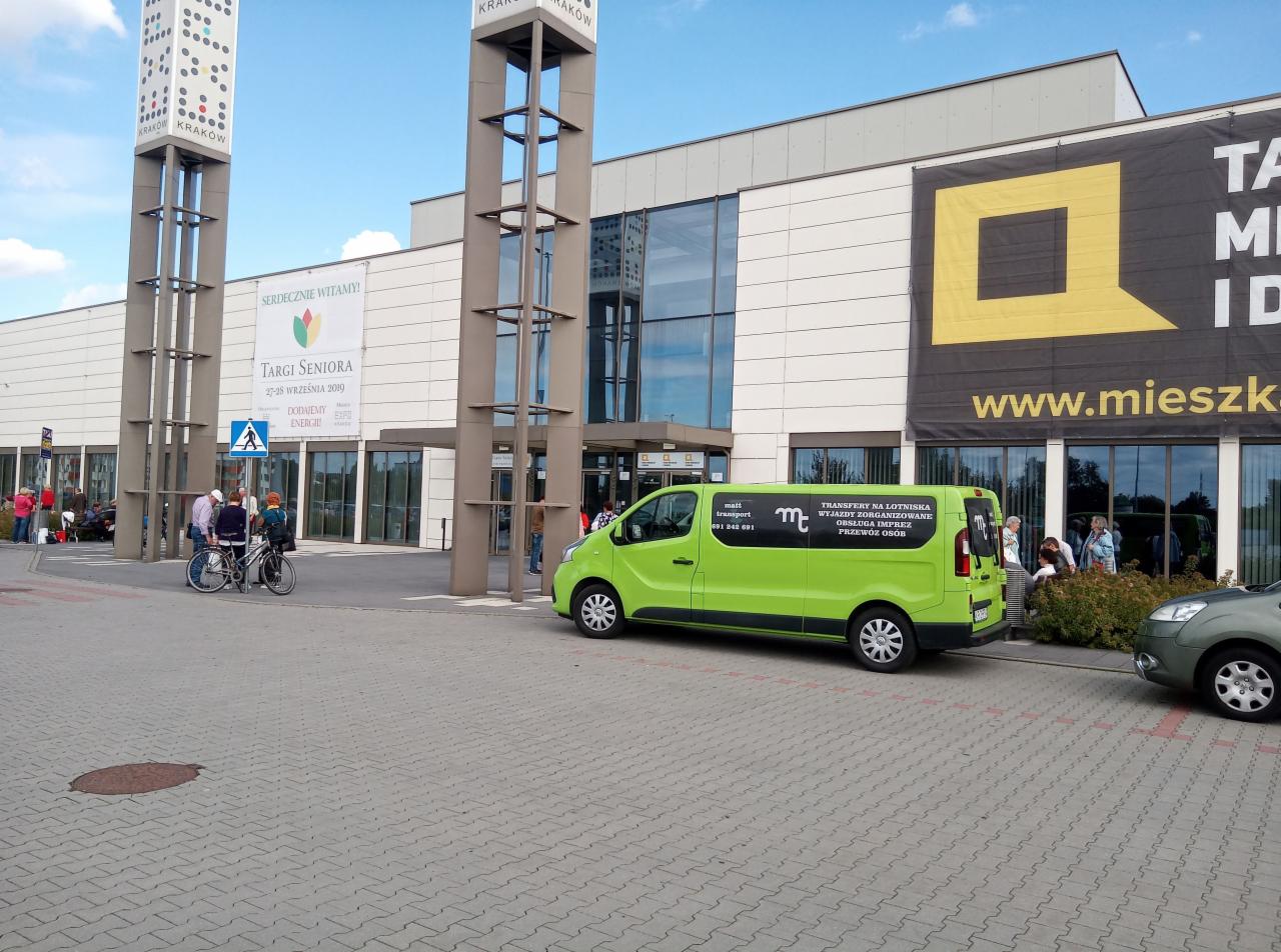 Jasnozielony bus z napisem 'Transfery na lotniska, wyjazdy zorganizowane, obsługa imprez, przewóz osób' zaparkowany przed budynkiem centrum targowego w Krakowie, na tle widoczny baner reklamowy.
