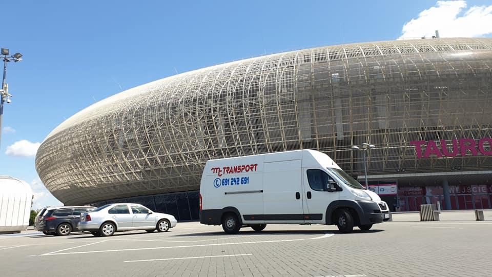 Biały bus z logo firmy transportowej na tle Stadionu Śląskiego w Rybniku, zaparkowany na parkingu.