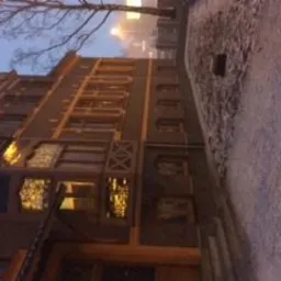 Fasada kamienicy z oknami oświetlonymi ciepłym światłem, widoczna pod kątem, z fragmentem chodnika i drzewa w tle, wieczorny krajobraz miejski.