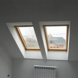 Dwa okna dachowe z drewnianymi ramami w białym, wykończonym pokoju na poddaszu, widok na sąsiednie domy w pochmurny dzień, biały grzejnik pod oknami.