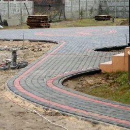 Kładzenie kostki brukowej: zakrzywiona ścieżka z czerwonym akcentem, obrzeża, palety kostki w tle, etap prac w ogrodzie.