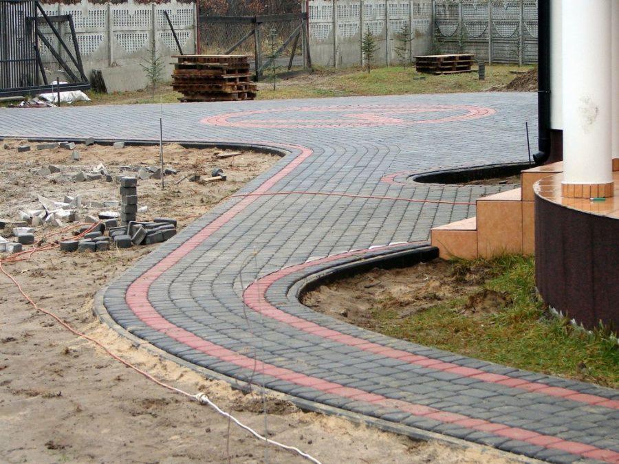 Kładzenie kostki brukowej: zakrzywiona ścieżka z czerwonym akcentem, obrzeża, palety kostki w tle, etap prac w ogrodzie.