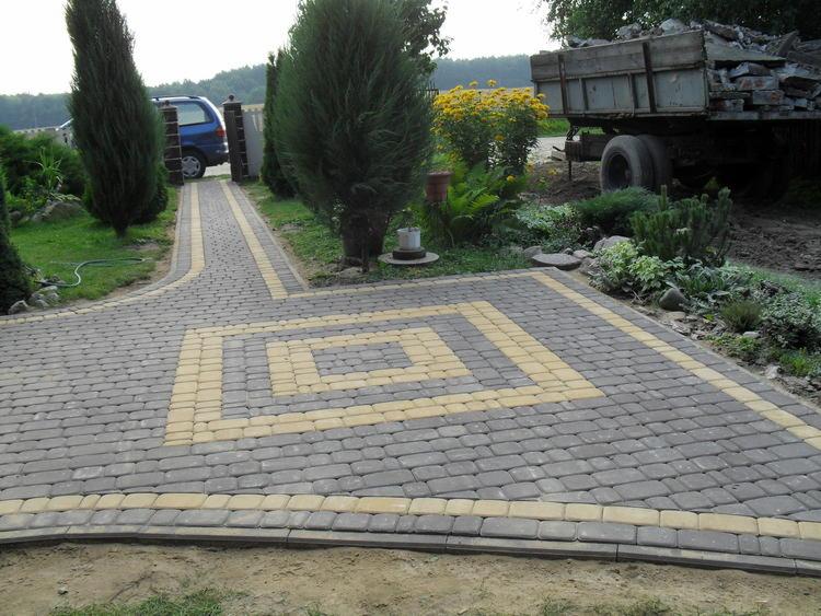 Ułożona kostka brukowa w odcieniach szarości i beżu tworzy geometryczny wzór na podjeździe. W tle widoczne rośliny, samochód i przyczepa z gruzem.