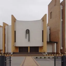 Nowoczesny kościół z aleją z kostki brukowej w odcieniach beżu i brązu, widzianą przez otwartą metalową bramę. Minimalistyczna elewacja, pochmurne niebo.