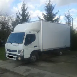 Biała ciężarówka dostawcza zaparkowana na podwórku, z widocznymi drzewami iglastymi i liściastymi w tle, w pochmurny dzień.