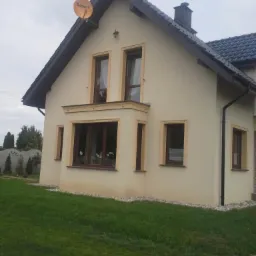 Elewacja domu jednorodzinnego w beżowym kolorze z ciemnymi ramami okiennymi i dachem. Widoczna antena satelitarna na szczycie dachu.