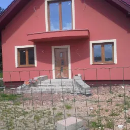 Czerwony dom jednorodzinny z brązowymi oknami i drzwiami. Widoczne schody wejściowe i elementy wykończeniowe elewacji. Budynek otoczony zielenią, ogrodzony siatką.