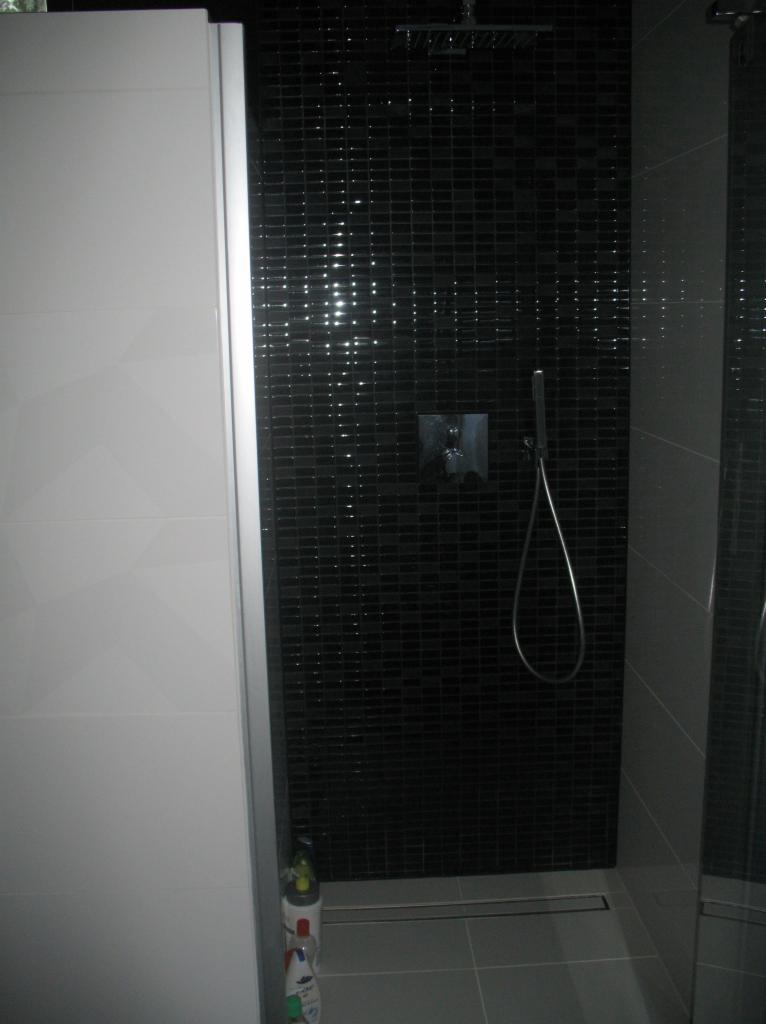 Kabina prysznicowa z czarną mozaiką i deszczownicą, biała ściana z geometrycznym wzorem, odpływ liniowy. Nowoczesny design łazienki.