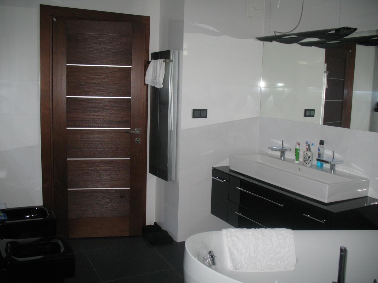 Nowoczesna łazienka z brązowymi drzwiami z poziomymi wstawkami, czarną armaturą i białą ceramiką. Widoczny fragment wanny i umywalki.