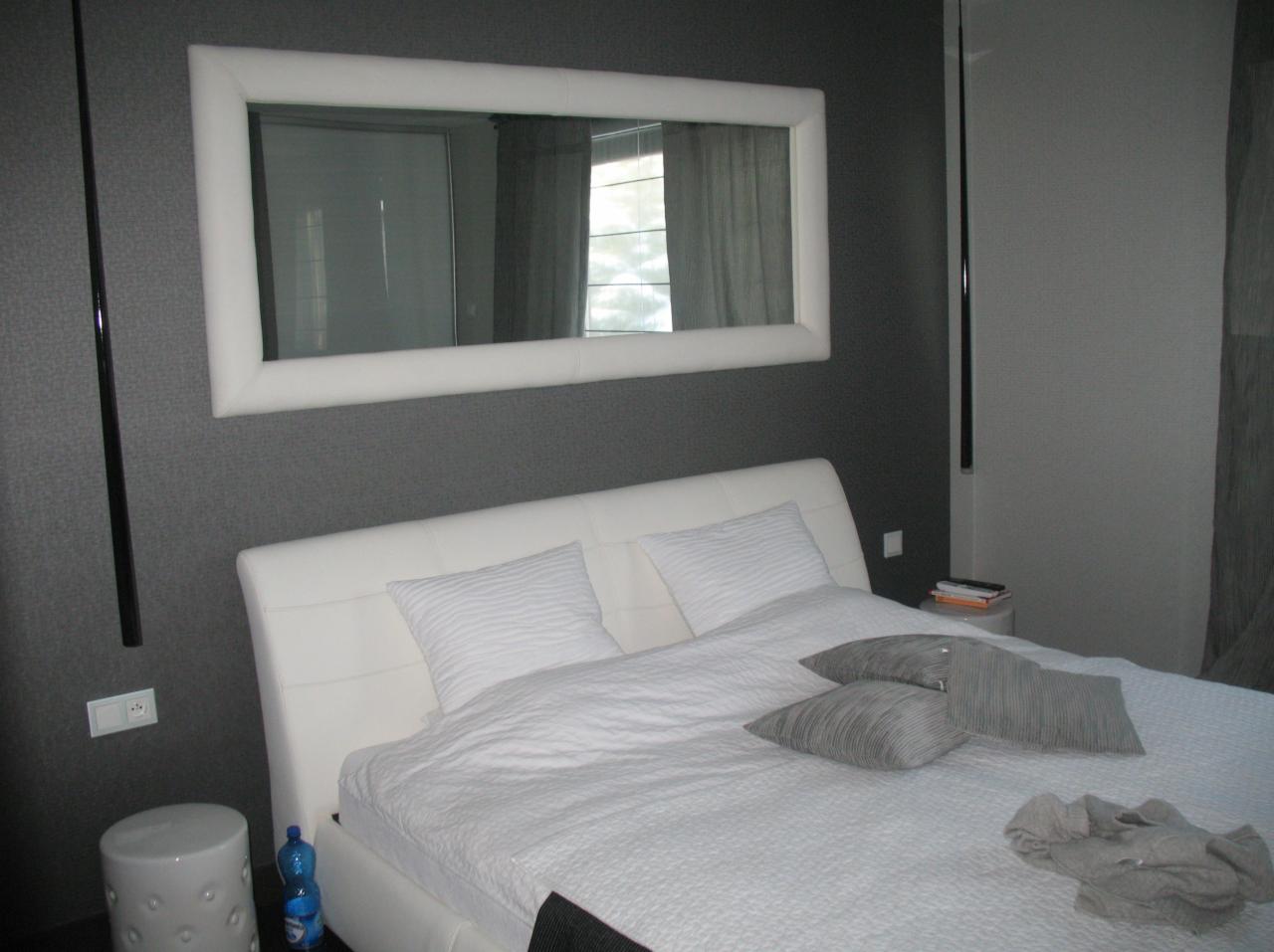 Elegancka sypialnia z białym łóżkiem, dużym lustrem w białej ramie na szarej ścianie i minimalistycznym oświetleniem. Nowoczesny design wnętrza.