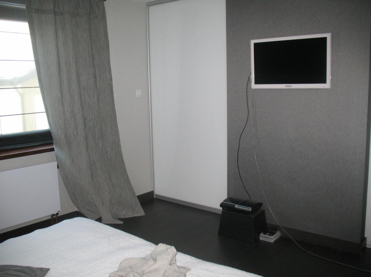 Minimalistyczna sypialnia w szarościach: białe łóżko, grafitowa ściana z TV, zasłona w paski i biała szafa przesuwna. Nowoczesny design.