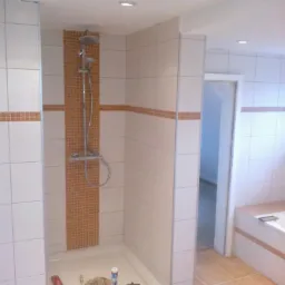 Wykończona kabina prysznicowa z mozaiką i białą armaturą, część wykończonej łazienki z beżowymi płytkami podłogowymi i wanną. Widoczne narzędzia glazurnicze.