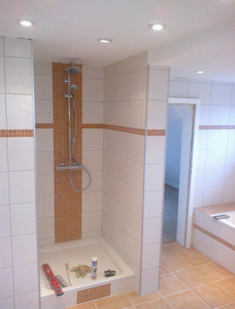 Wykończona kabina prysznicowa z mozaiką i białą armaturą, część wykończonej łazienki z beżowymi płytkami podłogowymi i wanną. Widoczne narzędzia glazurnicze.