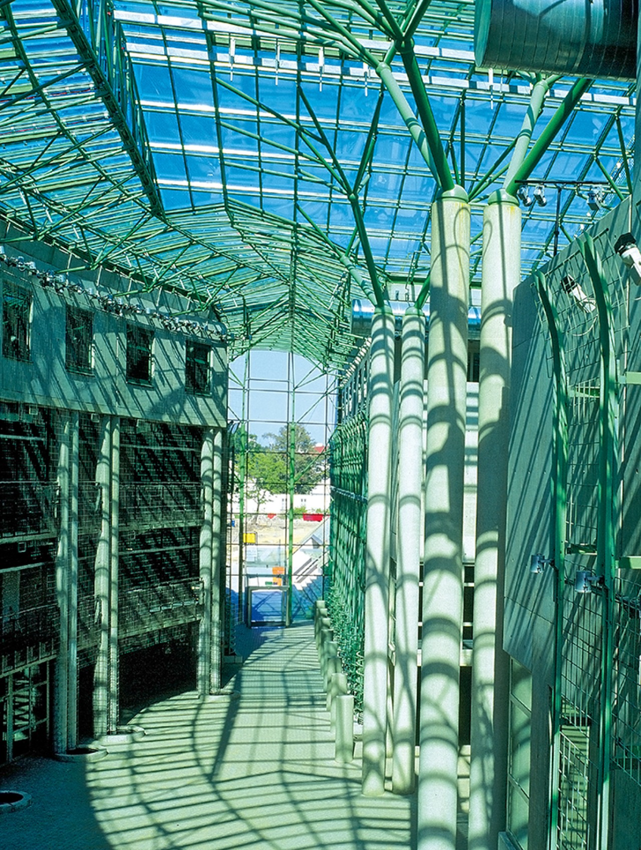 Wnętrze budynku z przeszklonym dachem i zieloną konstrukcją nośną. Widoczne kolumny i gra świateł. Architektura industrialna z elementami nowoczesności.