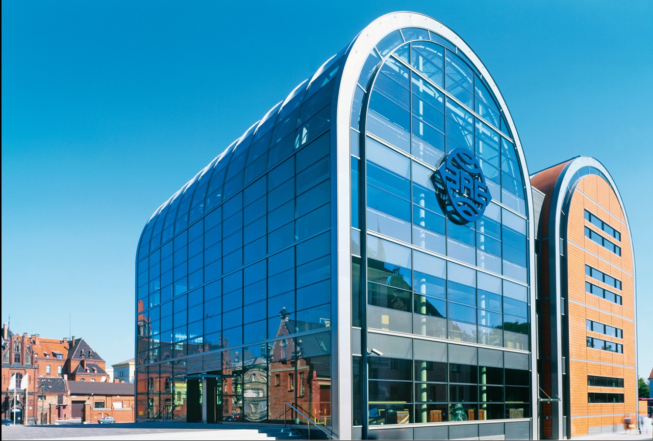 Nowoczesny budynek biurowy z przeszkloną fasadą w kształcie łuku, z logo firmy na elewacji. W tle widoczne starsze budynki i błękitne niebo.