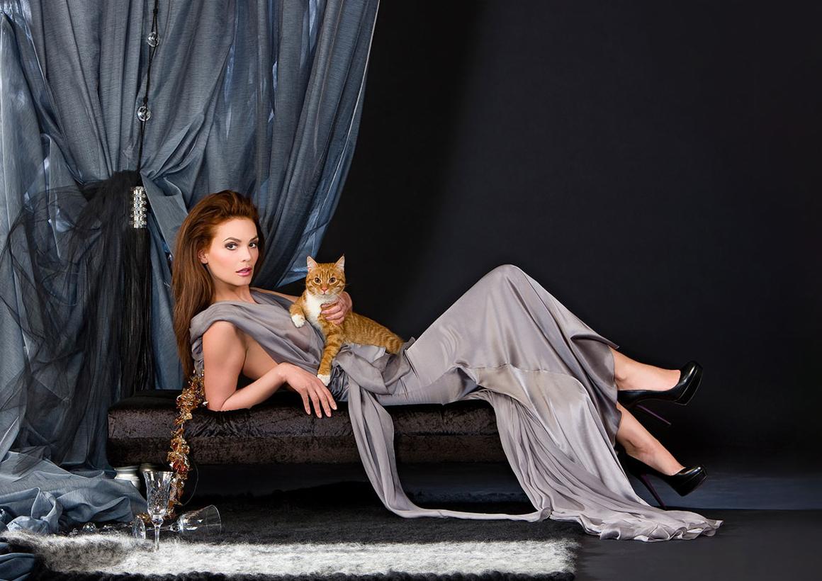 Kobieta w szarej sukni pozuje z rudym kotem na tle czarnego studia z drapowanymi tkaninami i rozbitymi kieliszkami