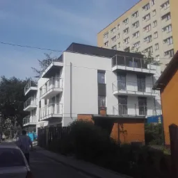 Nowoczesny budynek wielorodzinny z białymi ścianami i balkonami, widok z ulicy, na drugim planie wysoki blok mieszkalny.