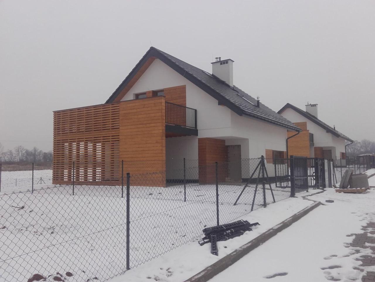 Nowoczesny dom jednorodzinny z drewnianą elewacją i balkonem, widoczny w zimowej scenerii z zasypanym śniegiem terenem i ogrodzeniem z siatki.