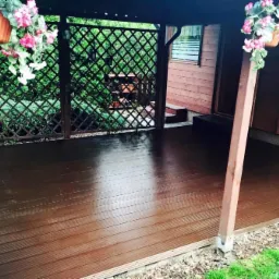 Zadaszony obszar z ciemnobrązową podłogą z desek, ozdobiony wiszącymi donicami z r&oacute;żowymi kwiatami i zieloną kratą w tle.