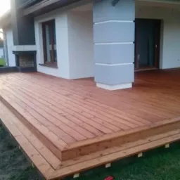 Nowoczesny taras z drewna egzotycznego z dwoma stopniami, przylegający do domu z elementami kamiennymi i kolumną, otoczony trawnikiem i niską zielenią.