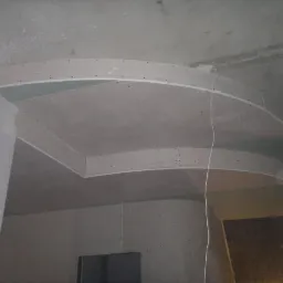 Sufit podwieszany z płyt gipsowo-kartonowych w kształcie łuku, widoczne mocowania i przewody. W tle okno i surowa ściana. Prace wykończeniowe w trakcie realizacji.