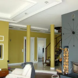 Salon po remoncie: żółte ściany, białe sztukaterie sufitowe, kolumny, szare wykończenie kominka z zegarem i drewnem, spiralne schody.