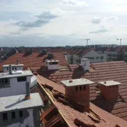 Nowe, czerwone dachówki ceramiczne układane na dachu budynku wielorodzinnego, widoczne kominy w trakcie budowy, w tle zabudowa miejska i żurawie budowlane.