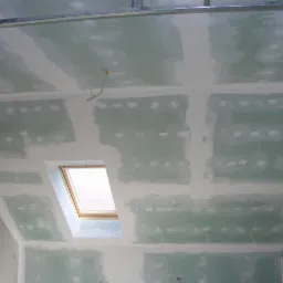 Sufit poddasza ze skosem i oknem dachowym po szpachlowaniu, widoczne płyty gipsowo-kartonowe i metalowa konstrukcja nośna.