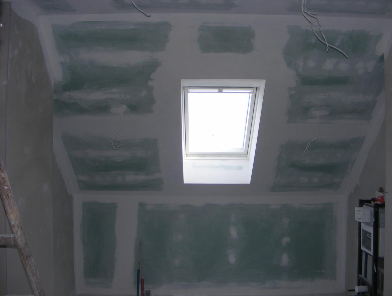 Poddasze ze ścianami w trakcie szpachlowania, widoczne fragmenty płyt gipsowo-kartonowych, okno dachowe i wiszące przewody elektryczne. Częściowo zagruntowane ściany.