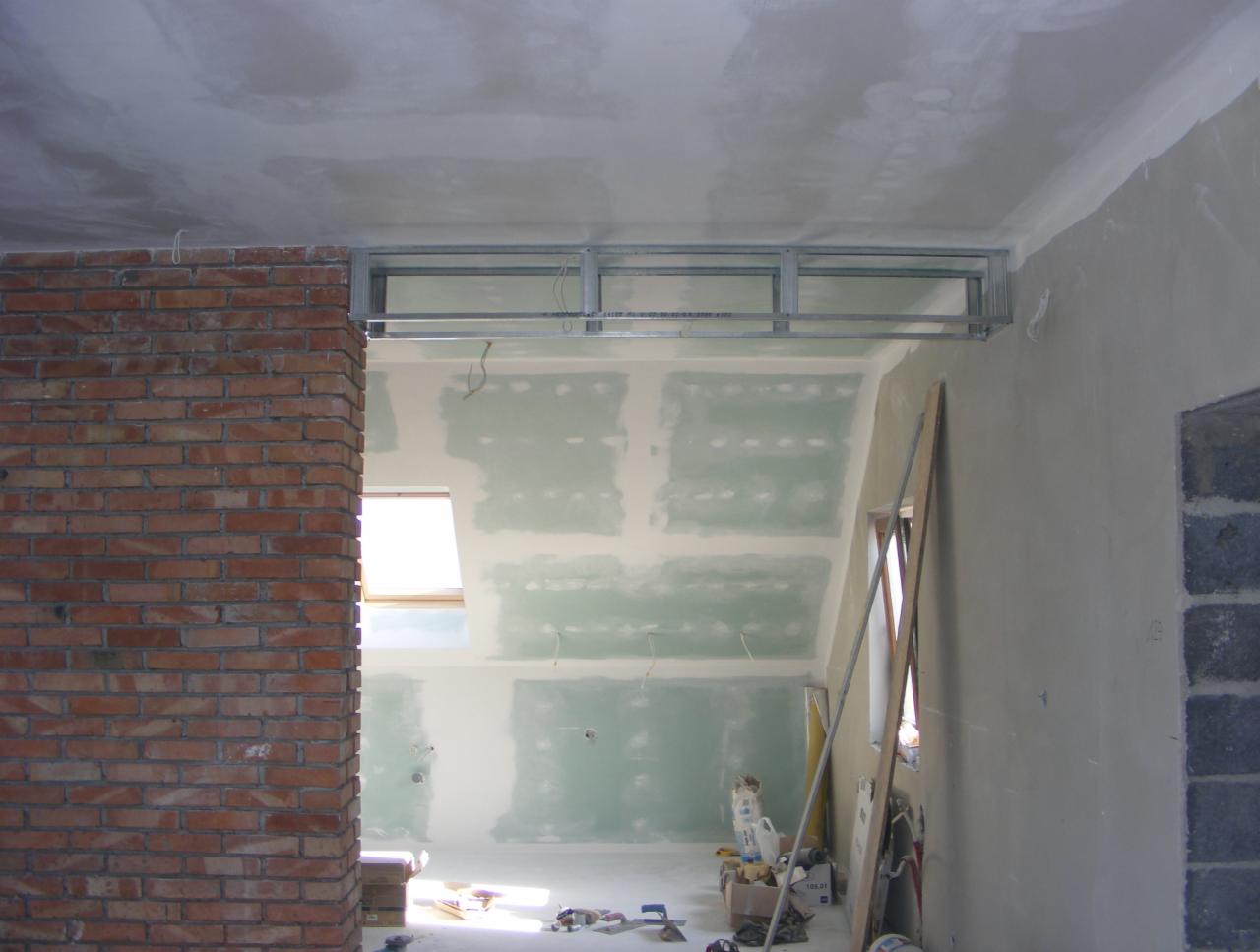 Wnętrze w trakcie prac wykończeniowych: ściany z płyt gipsowych, konstrukcja sufitu podwieszanego, fragment ściany z czerwonej cegły, narzędzia pracy na podłodze.