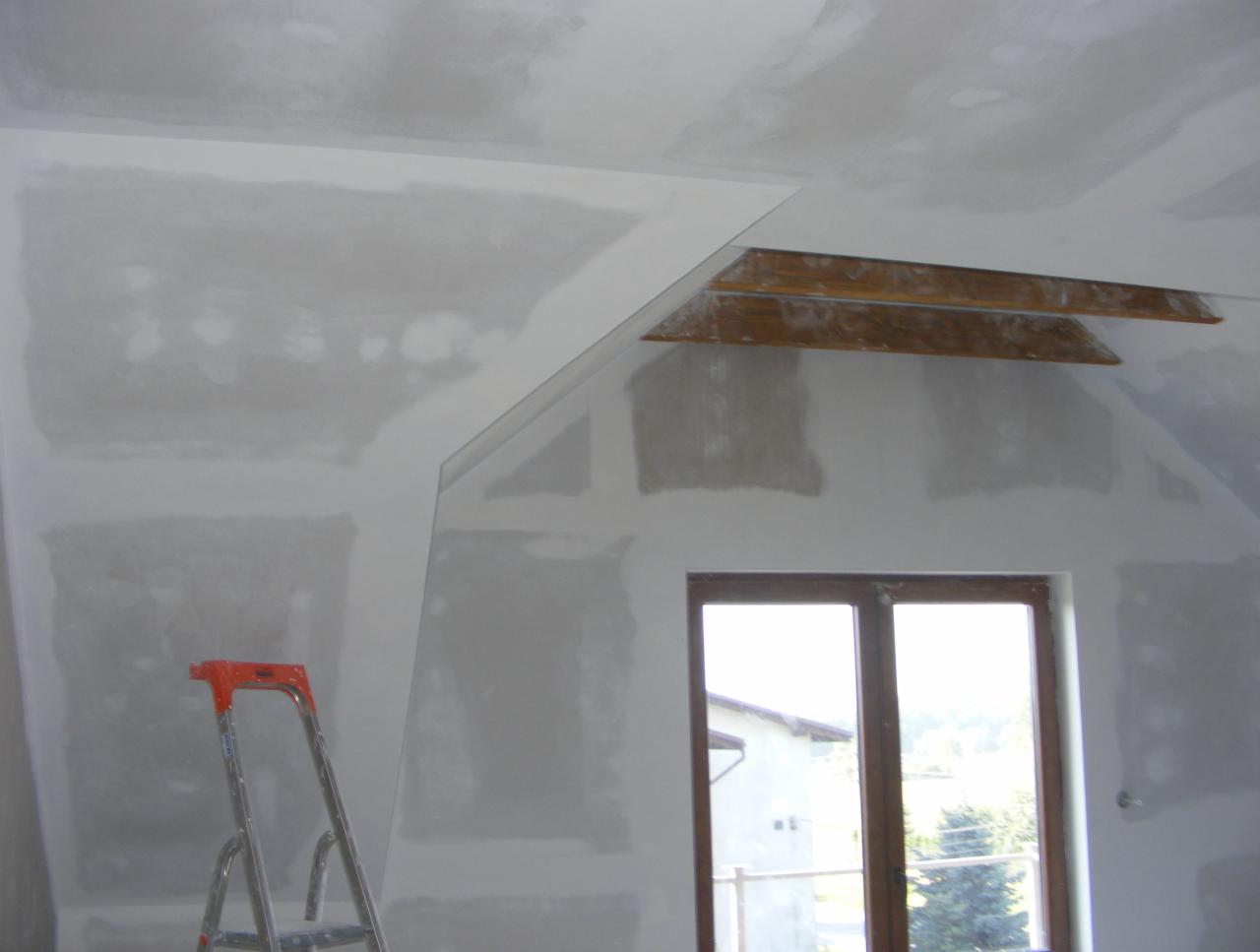 Poddasze po szpachlowaniu, widoczna drabina i okno. Ściany przygotowane do malowania, z widocznymi drewnianymi elementami konstrukcyjnymi.
